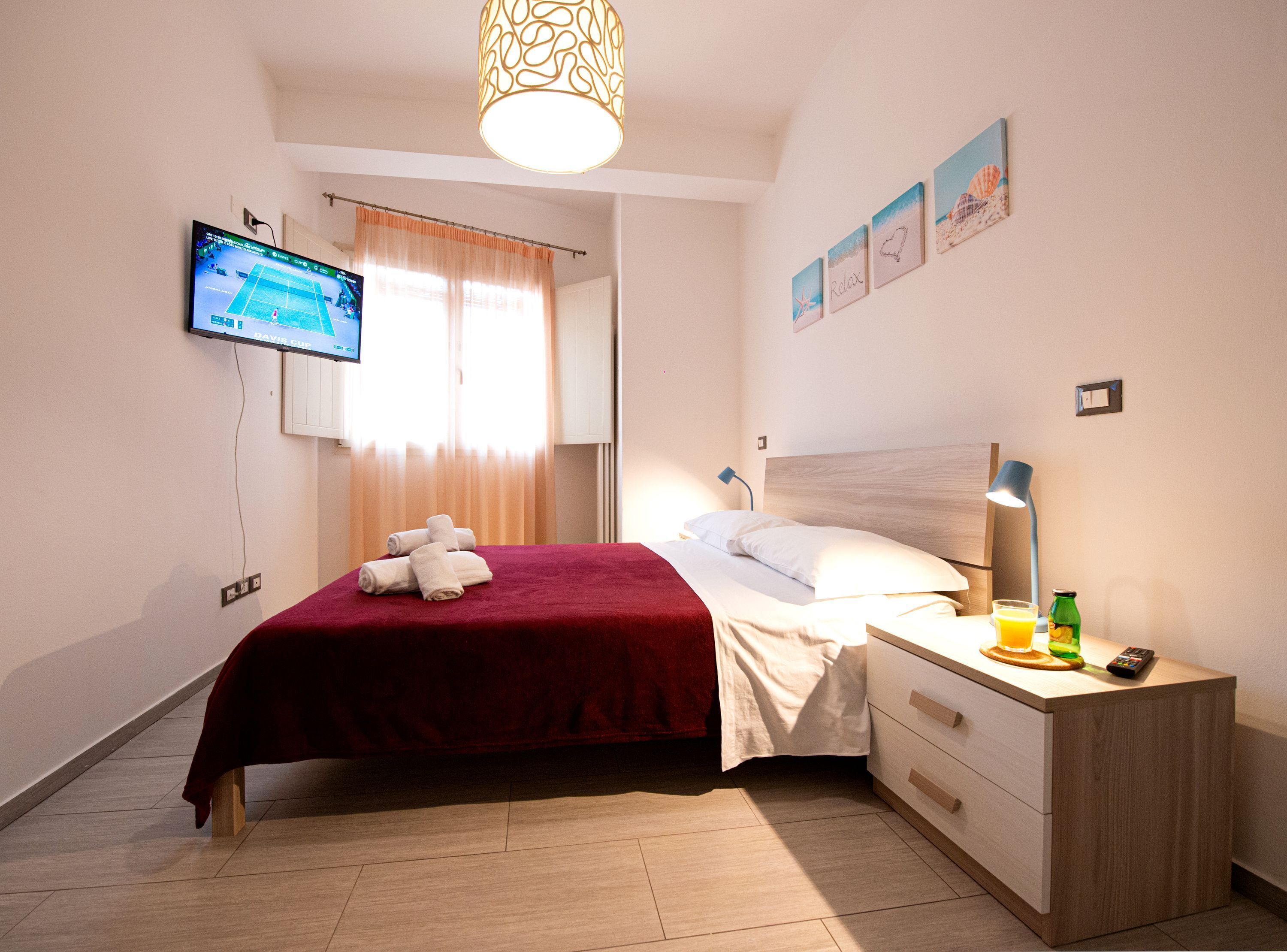 Una habitación de hotel con cama, mesita de noche con lámpara, televisión en la pared y ventana con cortinas.
