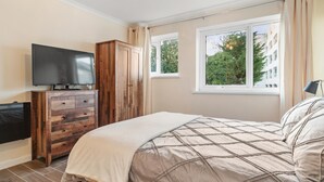 Une chambre à coucher comprenant un lit, une commode en bois, un téléviseur et une fenêtre donnant sur des arbres et des immeubles.