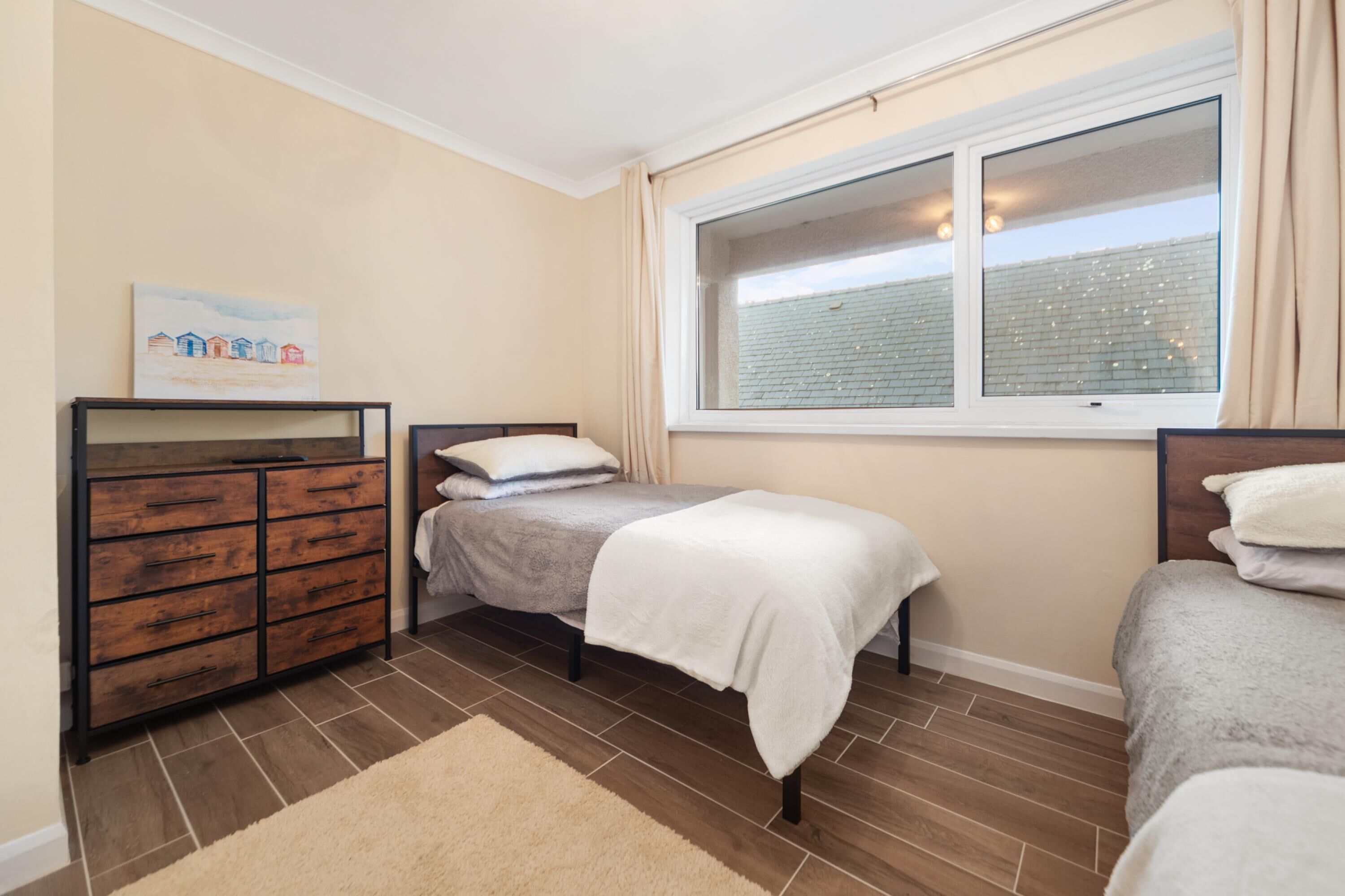 A bedroom with two beds, a wooden dresser, a window with curtains, and a painting on the wall.