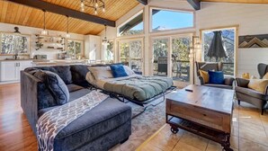 A spacious living area with a sofa, a bed, and a coffee table. There are large windows providing natural light and views of the outdoors. - Timber Peak Cabin! Hot Tub,BBQ & Scenic Views! (Divide)