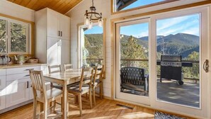 A wooden interior with a dining table and chairs, a kitchen with white cabinets, and a view of mountains through large sliding doors. - Timber Peak Cabin! Hot Tub,BBQ & Scenic Views! (Divide)