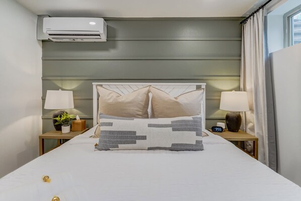 A hotel room with a bed, two bedside tables, a nightstand, an air conditioning unit, a clock, and a window with curtains.