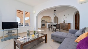 A living room with a TV, coffee table, and sofa, an open kitchen with dining area, and a balcony with outdoor furniture. - Wonderful and comfortable villa  with private pool in Benitachell, Costa Blanca, Spain for 6 persons (Benitachell)