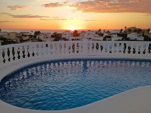 Une piscine bordée d’une balustrade blanche, entourée de bâtiments à l’architecture méditerranéenne, au coucher du soleil.
