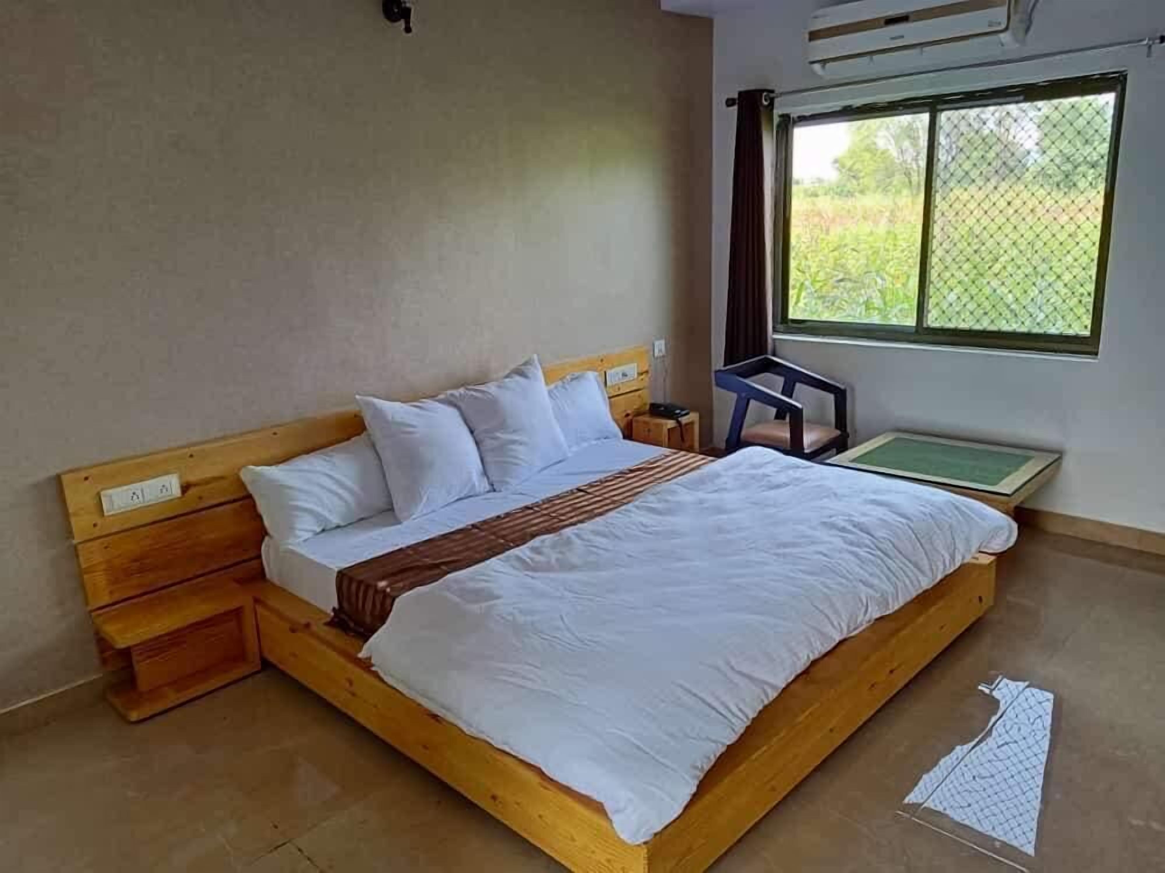 Une chambre avec un lit simple, une tête de lit en bois, du linge de lit blanc, une petite table de chevet et une fenêtre donnant sur un paysage verdoyant.