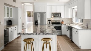 A modern kitchen with white cabinets, a marble countertop, stainless steel appliances, and two gold bar stools. - Private Pickleball Court, Movie Room, & Hot Tub! (Scottsdale)