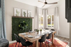 Un coin repas avec une table en bois dressée pour six personnes, entourée de chaises noires, d’un ventilateur de plafond et d’une composition de cactus fixée au mur.