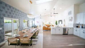 A spacious kitchen with white cabinets, a dining area with a long wooden table, and a bright living room with a sofa and a large window.