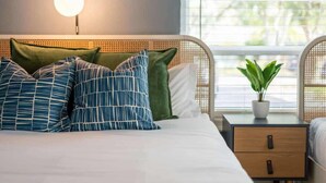 A bedroom with a bed, a bedside table with a plant, a wall-mounted light, and a window with a view of greenery.