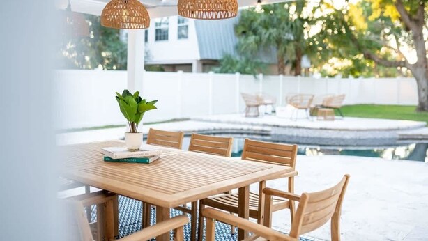An outdoor patio with a wooden table and chairs, surrounded by a white fence and a pool.
