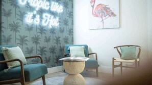 A room with a neon sign reading "Tropic like it's Hot," a tropical wallpaper, and a flamingo painting. There are two chairs and a round table with a book on it.