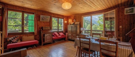 A wooden cabin interior with a dining table, chairs, a sofa, and large windows offering a view of greenery.