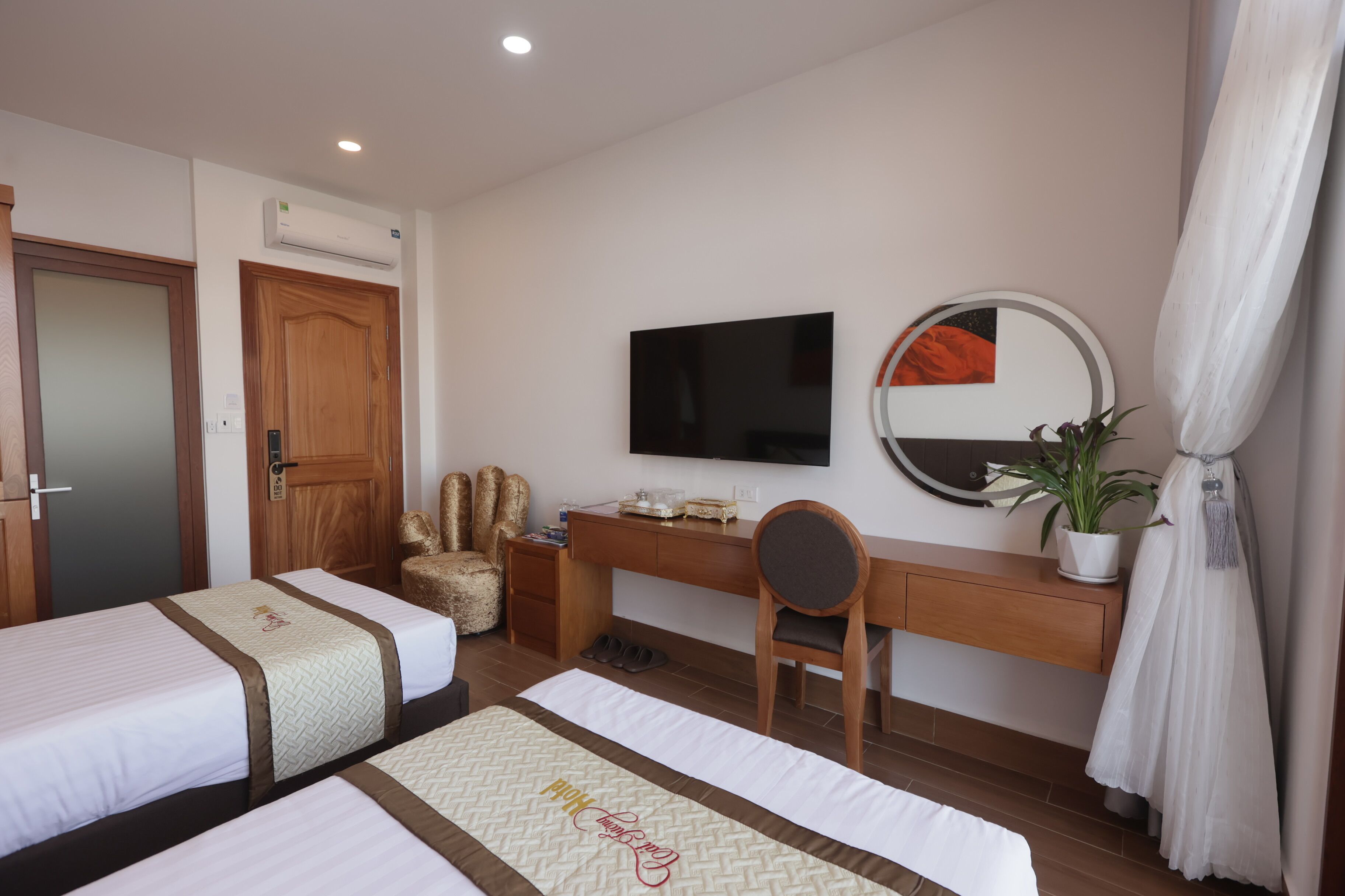A hotel room with two beds, a desk, a chair, a TV, a mirror, and a potted plant.