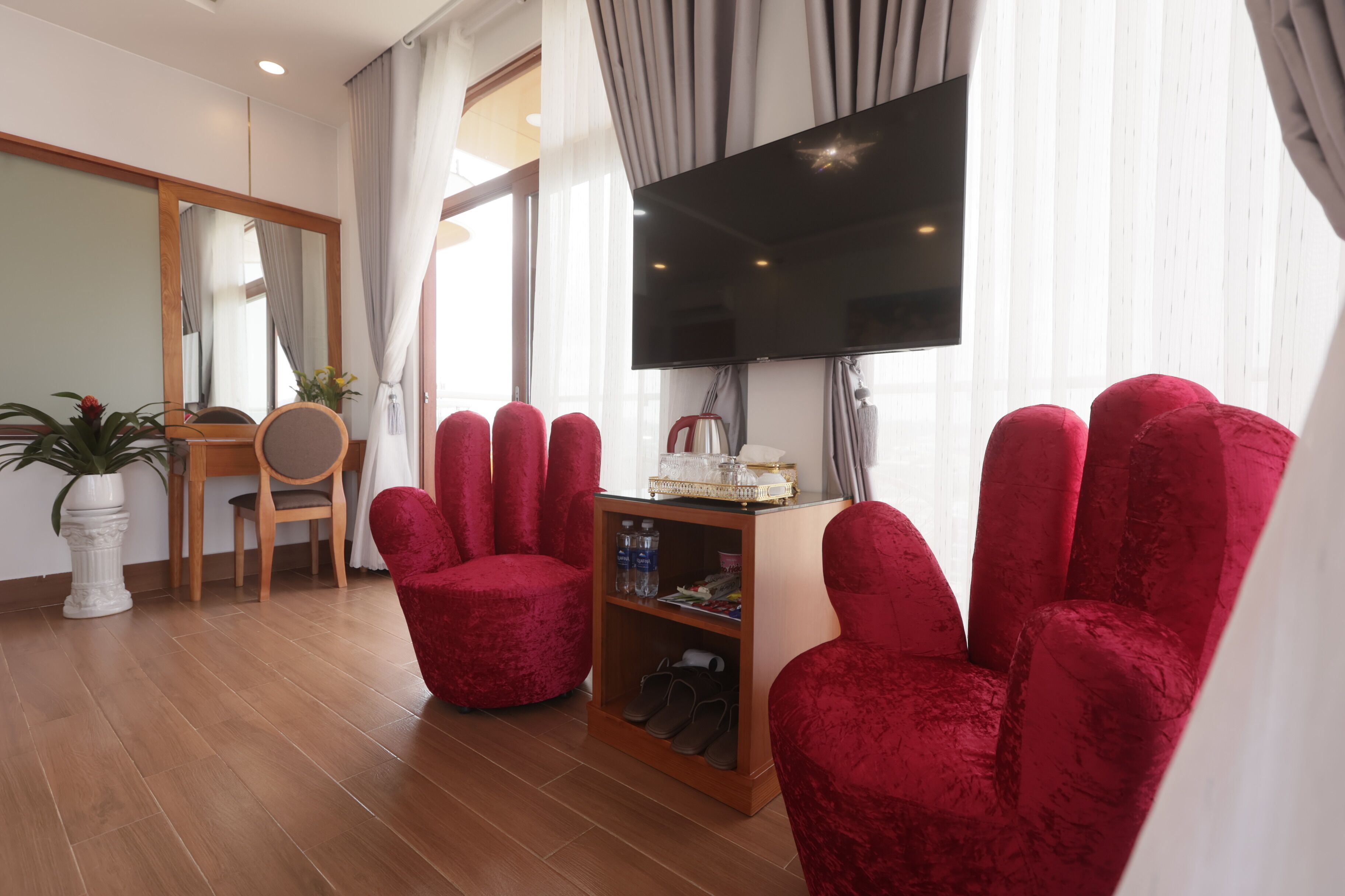 A modern living room with a flat-screen TV, red velvet armchairs, a wooden coffee table, and a small bookshelf.