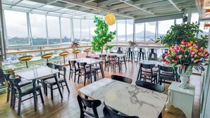 A restaurant with a high ceiling and large windows offering a view of the city and mountains. Tables and chairs are arranged for dining. - Cat Tuong Hotel (Lam Vien - Da Lat)