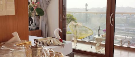A hotel room with a balcony, a bed with decorative pillows, a swan-shaped pillow, a glass table with a vase of flowers, and a view of the cityscape.