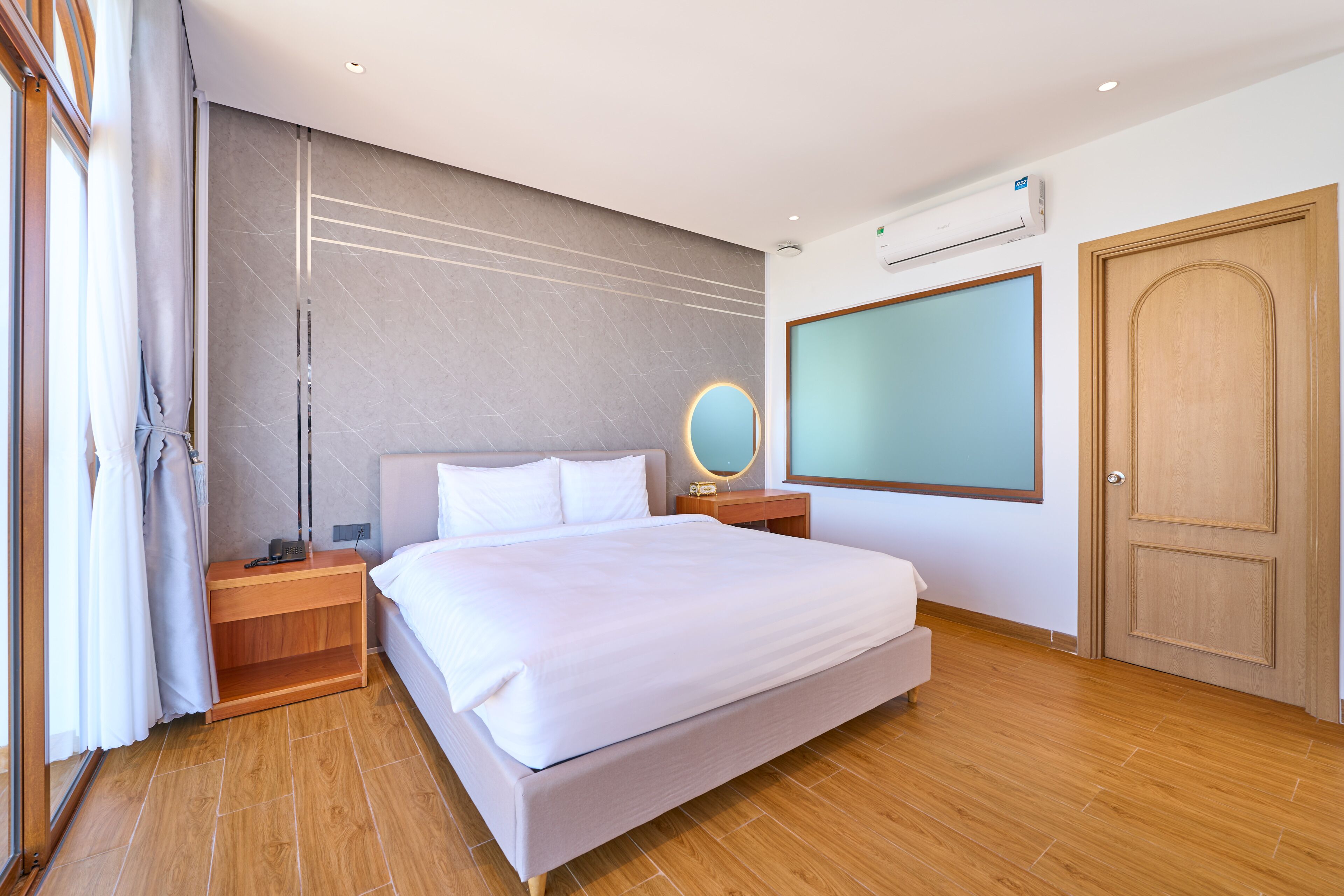 Una habitación de hotel moderna con una cama grande, mesitas de noche de madera, una mesilla con lámpara, un espejo en la pared y un amplio ventanal con cortinas.