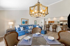 Ein modernes Wohnzimmer mit blauem Sofa, Esstisch mit Platzsets und einer Küchenzeile mit Barhocker.