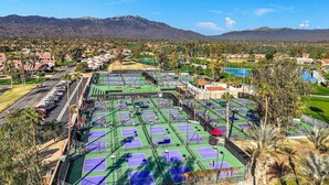 A tennis court complex with multiple courts, surrounded by residential buildings and a mountainous backdrop.