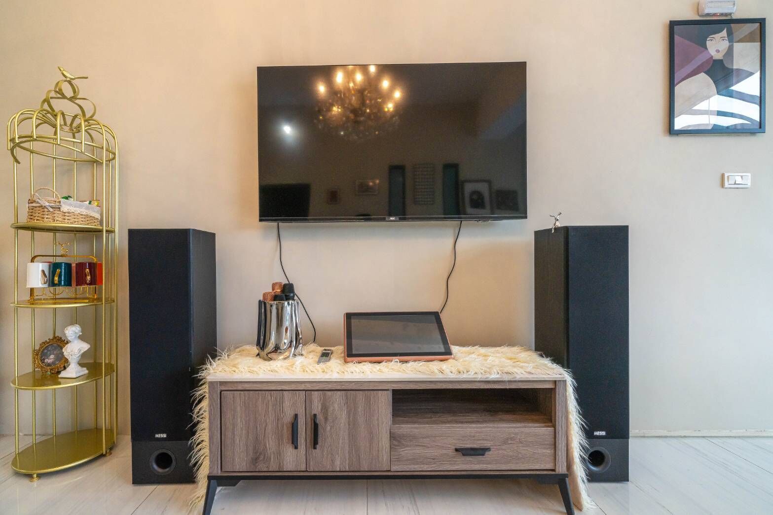 Ein modernes Wohnzimmer mit einem Wand-Fernseher, einer Holzkombination mit Schubladen, einem goldenen Metallregal und zwei schwarzen Lautsprechern.