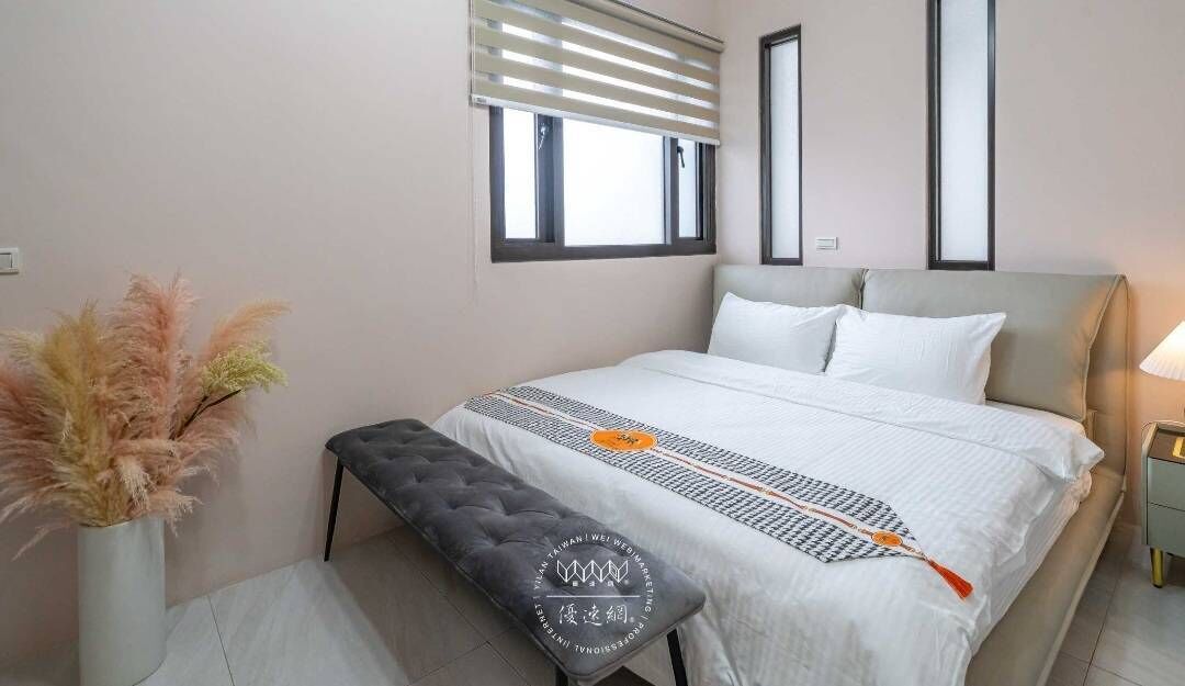 Un dormitorio con cama, mesitas de noche, un banco, una ventana con persianas y un jarrón con flores secas.