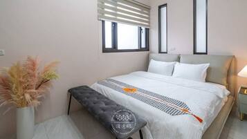 Un dormitorio con cama, mesitas de noche, un banco, una ventana con persianas y un jarrón con flores secas.