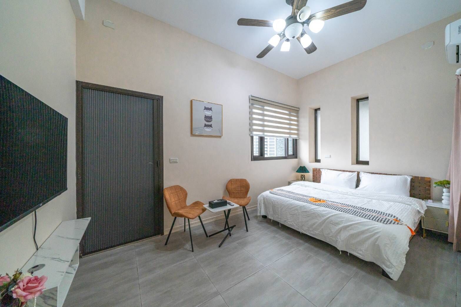 A bedroom with a bed, two chairs, a desk, a ceiling fan, a window with blinds, and a TV on the wall.