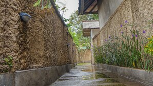 Un étroit sentier bordé de murs de terre et d’un canal d’eau, menant à un bâtiment traditionnel doté d’un toit en tuiles.