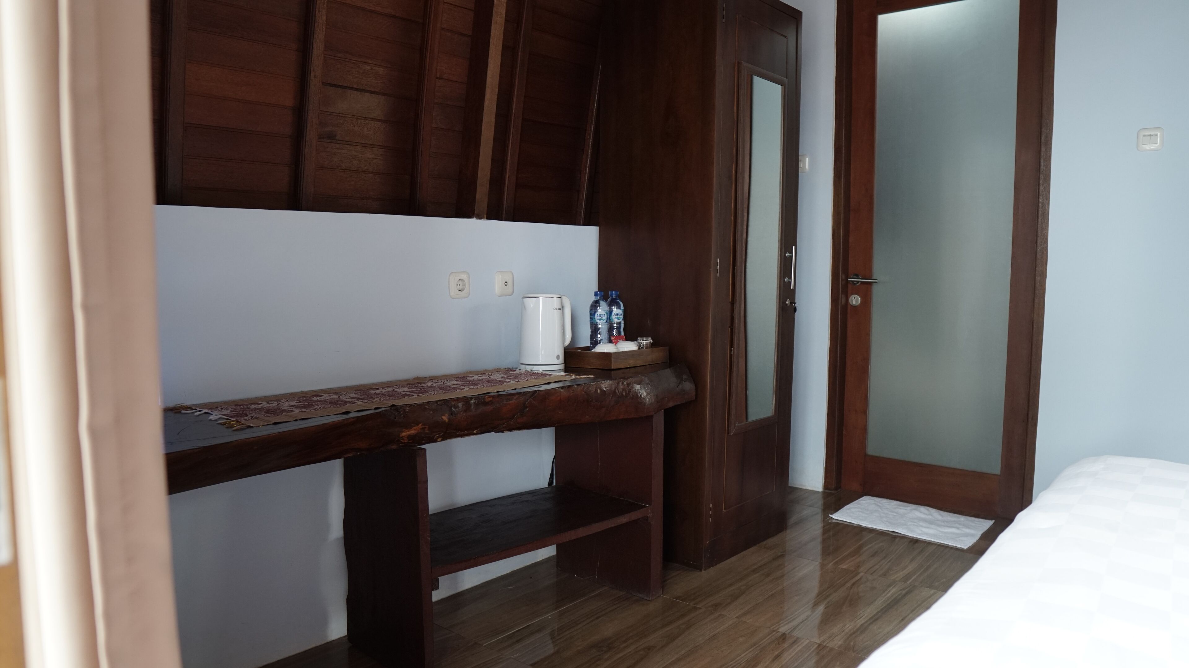 A room with a wooden bench, a small table with a coffee maker and water bottles, a bed, and a door with frosted glass panels.