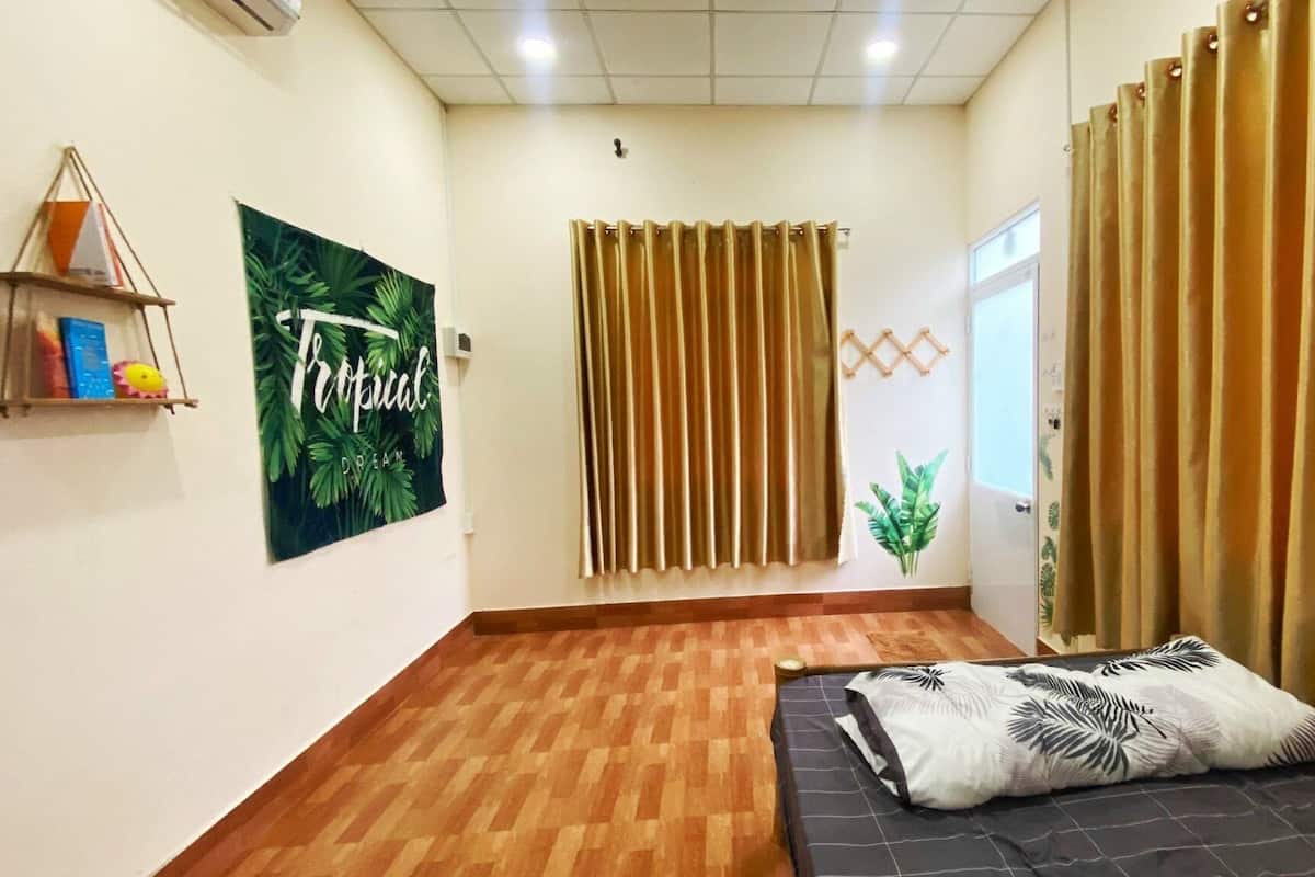 A hotel room with a wooden floor, a bed with a tropical-themed pillow, a wall with a 'Tropical Dream' poster, and a window with vertical blinds.