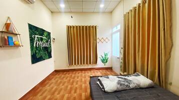 A hotel room with a wooden floor, a bed with a tropical-themed pillow, a wall with a 'Tropical Dream' poster, and a window with vertical blinds.