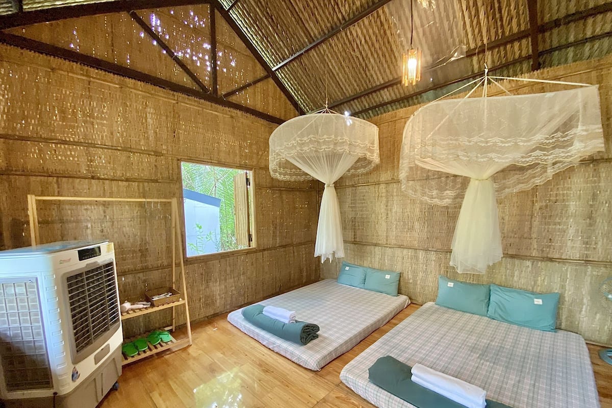 A room with two beds, a ceiling fan, and a window with a view of greenery.