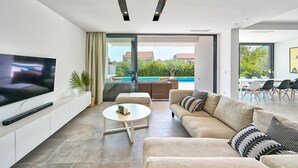 A modern living room with a flat-screen TV, a round coffee table, a beige sofa, and a glass door leading to a pool area.