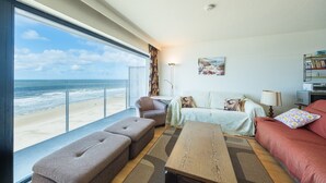 Ein geräumiges Wohnzimmer mit großem Fenster mit Blick auf einen Strand, einer Couch, einem Couchtisch und einem Bücherregal.