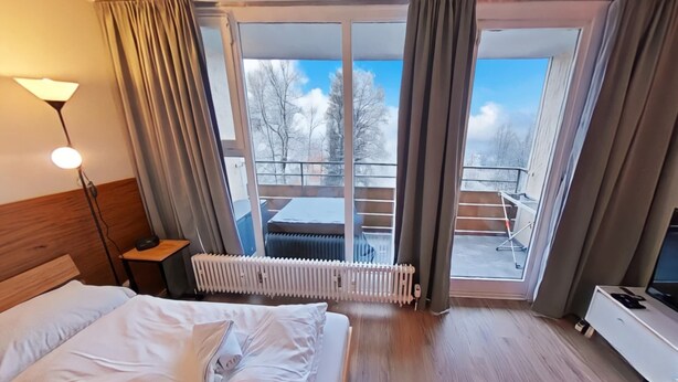 Ett hotellrum med en säng, en sänglampa, ett element, en tv och en balkong med utsikt över träd och en klar himmel.