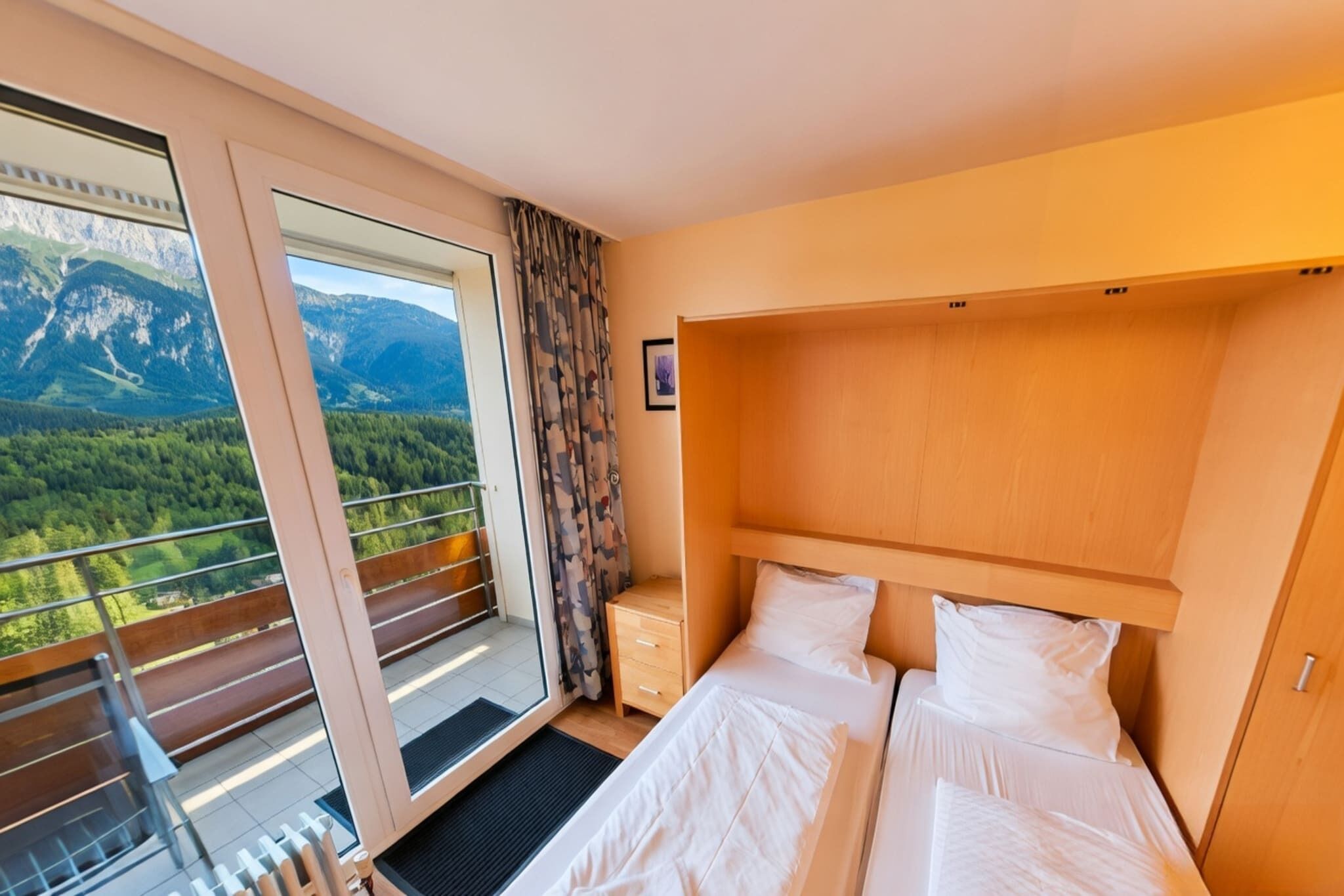 A hotel room with a bed, a nightstand, a wooden headboard, a window with a mountain view, and a balcony with a railing.