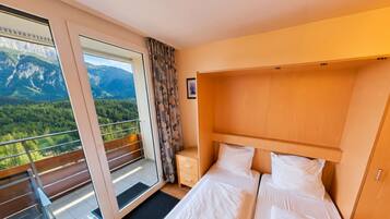 Ein Hotelzimmer mit Bett, Nachttisch, Holz-Kopfteil, Fenster mit Bergblick und Balkon mit Geländer.