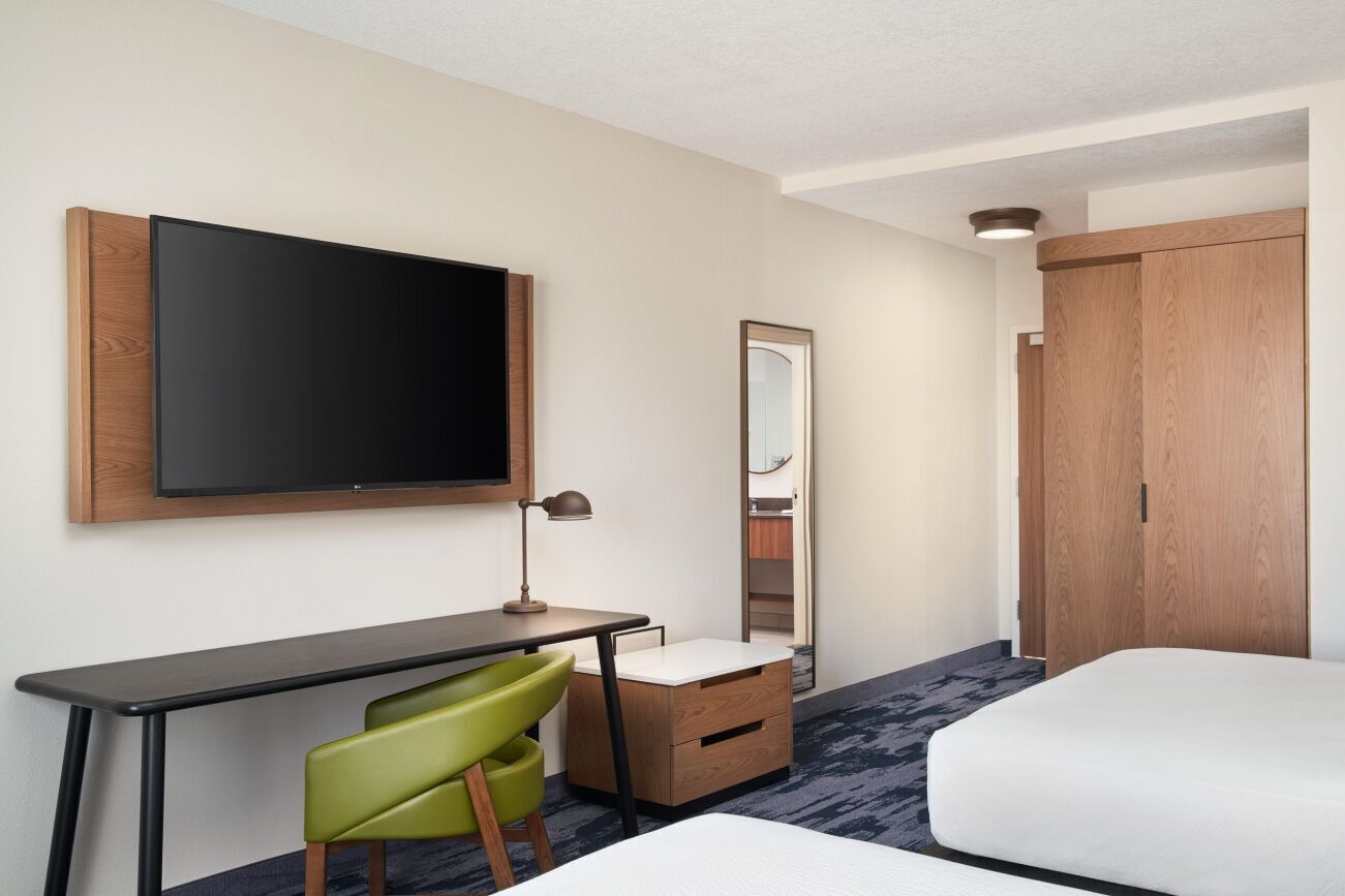 A hotel room with a bed, a desk with a chair, a flat-screen TV, a mirror, and a wardrobe.