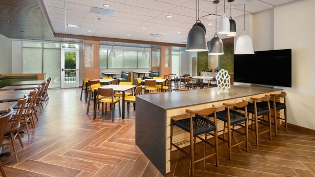 A modern dining area with a central island, multiple seating options, and a large flat-screen TV on the wall. - NEW - Fairfield Inn & Suites by Marriott Orlando at Millenia (Orlando)