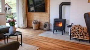 A cozy living room with a wooden floor, a TV mounted on the wall, a fireplace, and a stack of firewood.