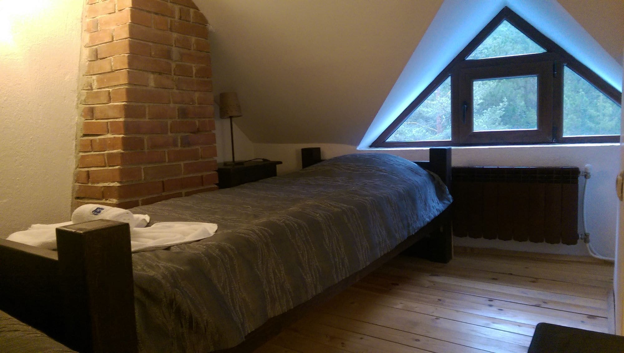 Una cama individual en una habitación con techo inclinado, pared de ladrillo y ventana de diseño triangular.