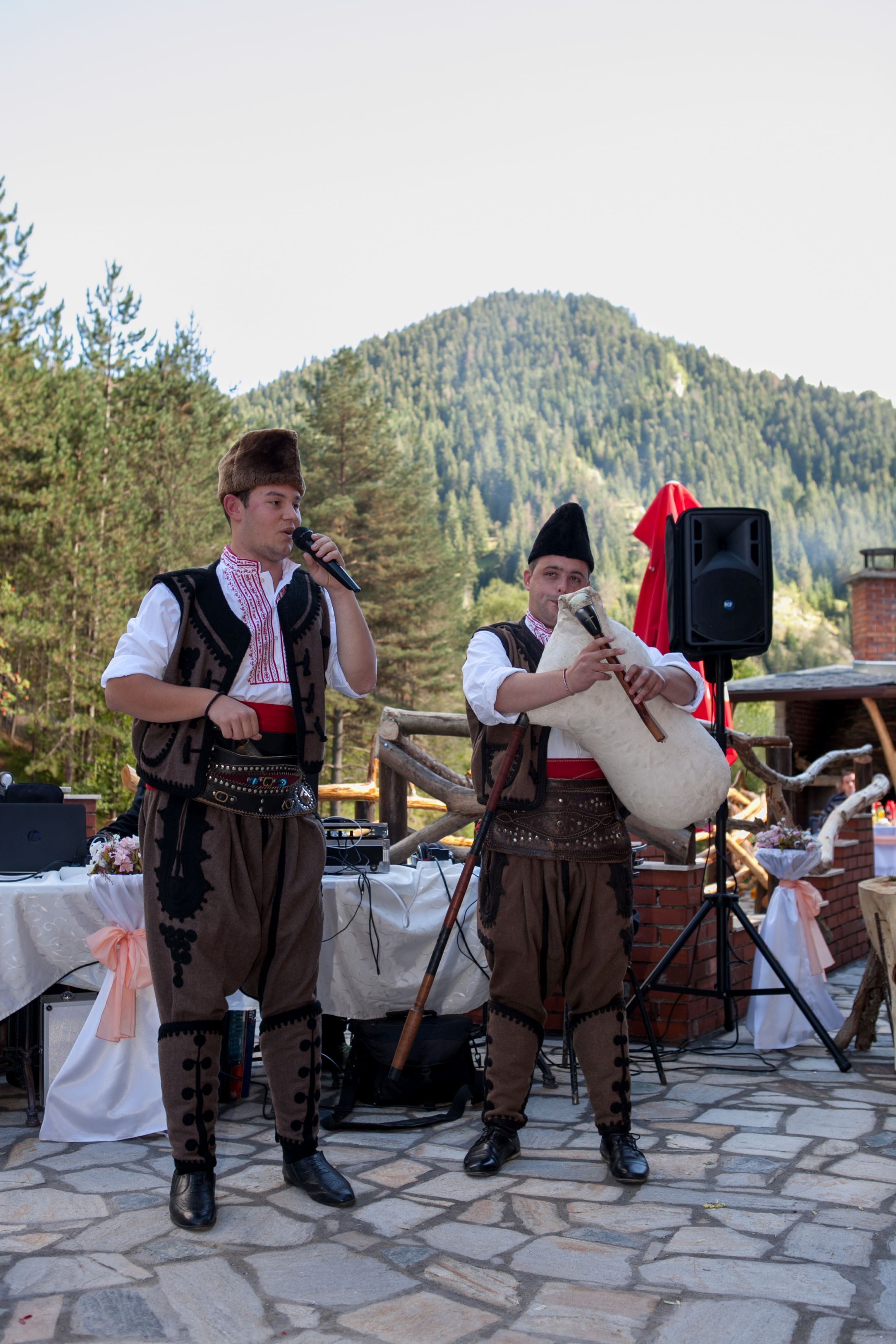 Two individuals in traditional attire performing outdoors with a mountainous backdrop.