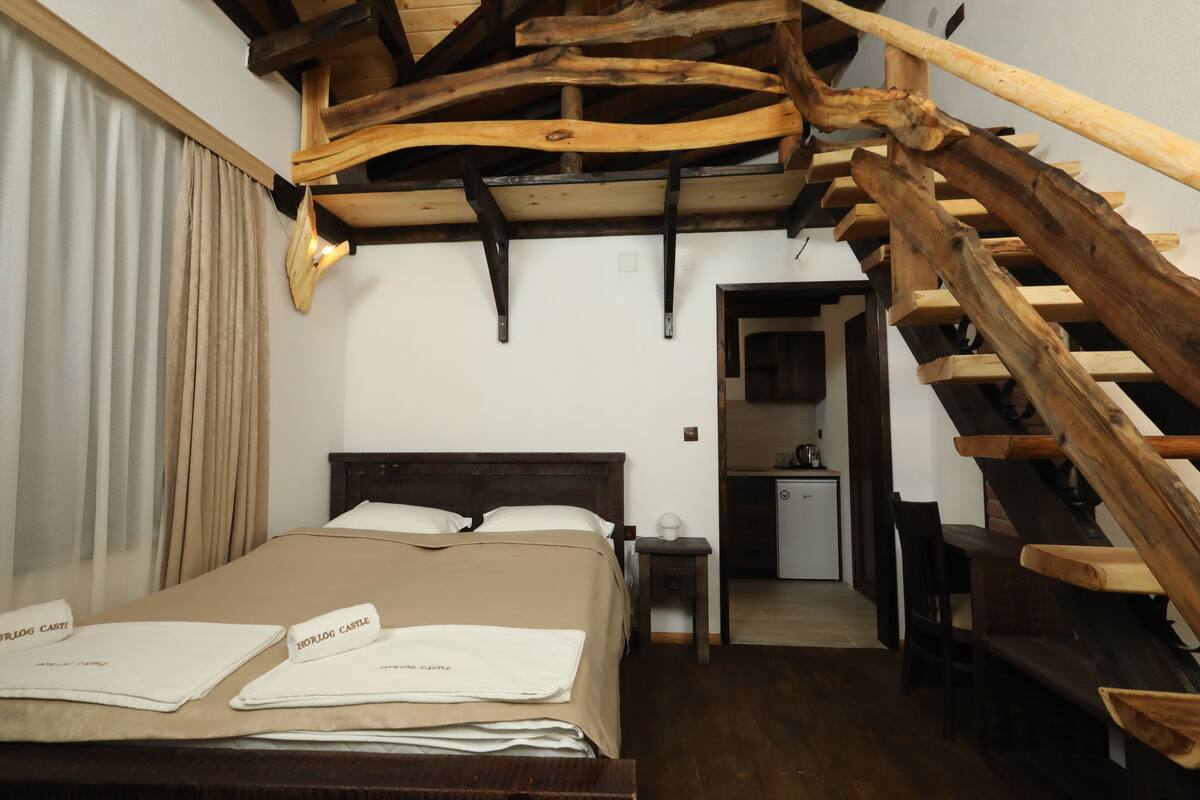 A hotel room with a wooden staircase, a bed with a beige bedspread, and a small table with a lamp.