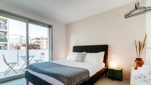 A modern hotel room with a large bed, a bedside table with a lamp, a view of the city through large windows, and a balcony with chairs. - Kristall 9  (Massagno)