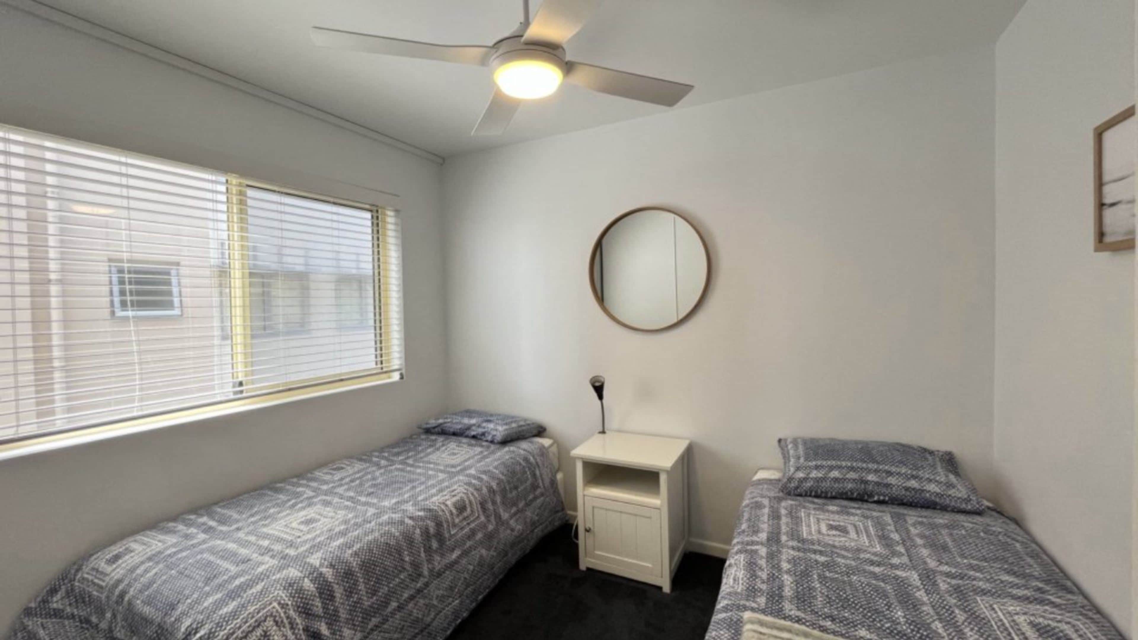 A bedroom with two single beds, a ceiling fan, a bedside table, and a round mirror.