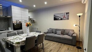 A modern kitchen with a dining area, a grey sofa, and a floor lamp. - PARADISE HOME (Augusta)