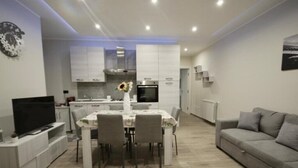 A modern living room with a grey sofa, a dining area with a table and chairs, and a kitchen with white cabinets and appliances. - PARADISE HOME (Augusta)