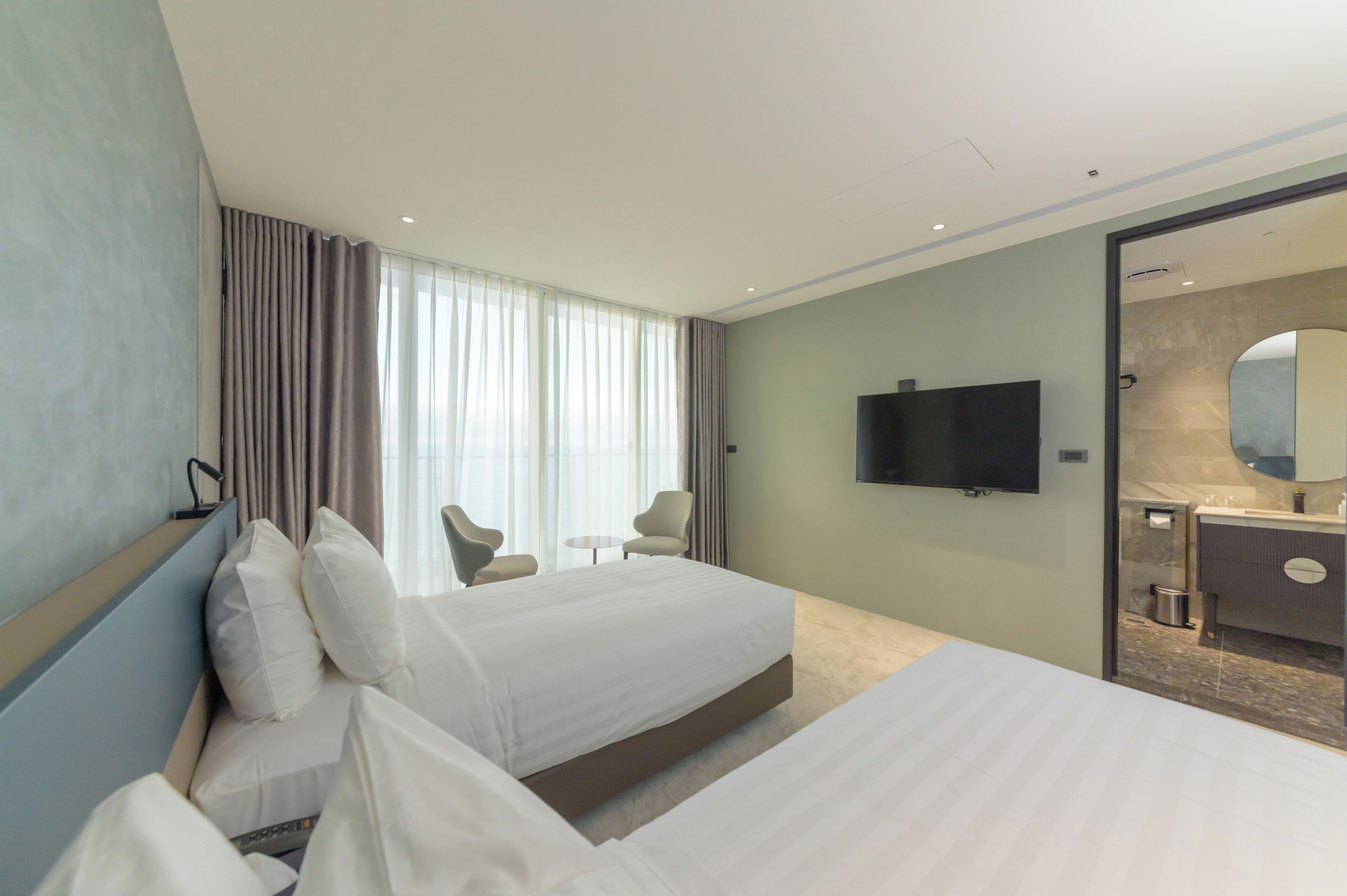 A hotel room with a large bed, two armchairs, a TV, and a bathroom visible through a glass door.