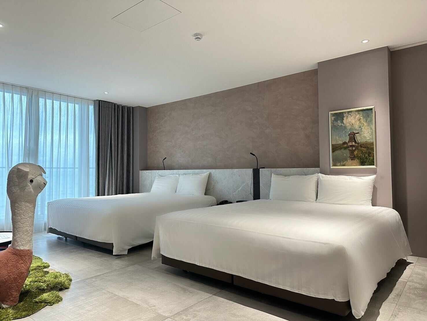 A modern hotel room with two beds, a large window with curtains, a painting on the wall, and a unique sculpture on the floor.