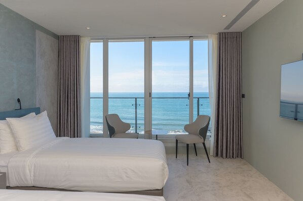 A hotel room with a large bed, two chairs, a TV, and a balcony with a view of the sea.